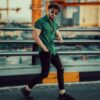 Stylish man with sunglasses in green shirt and ripped jeans posing on bridge against urban backdrop.