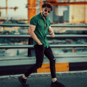 Stylish man with sunglasses in green shirt and ripped jeans posing on bridge against urban backdrop.