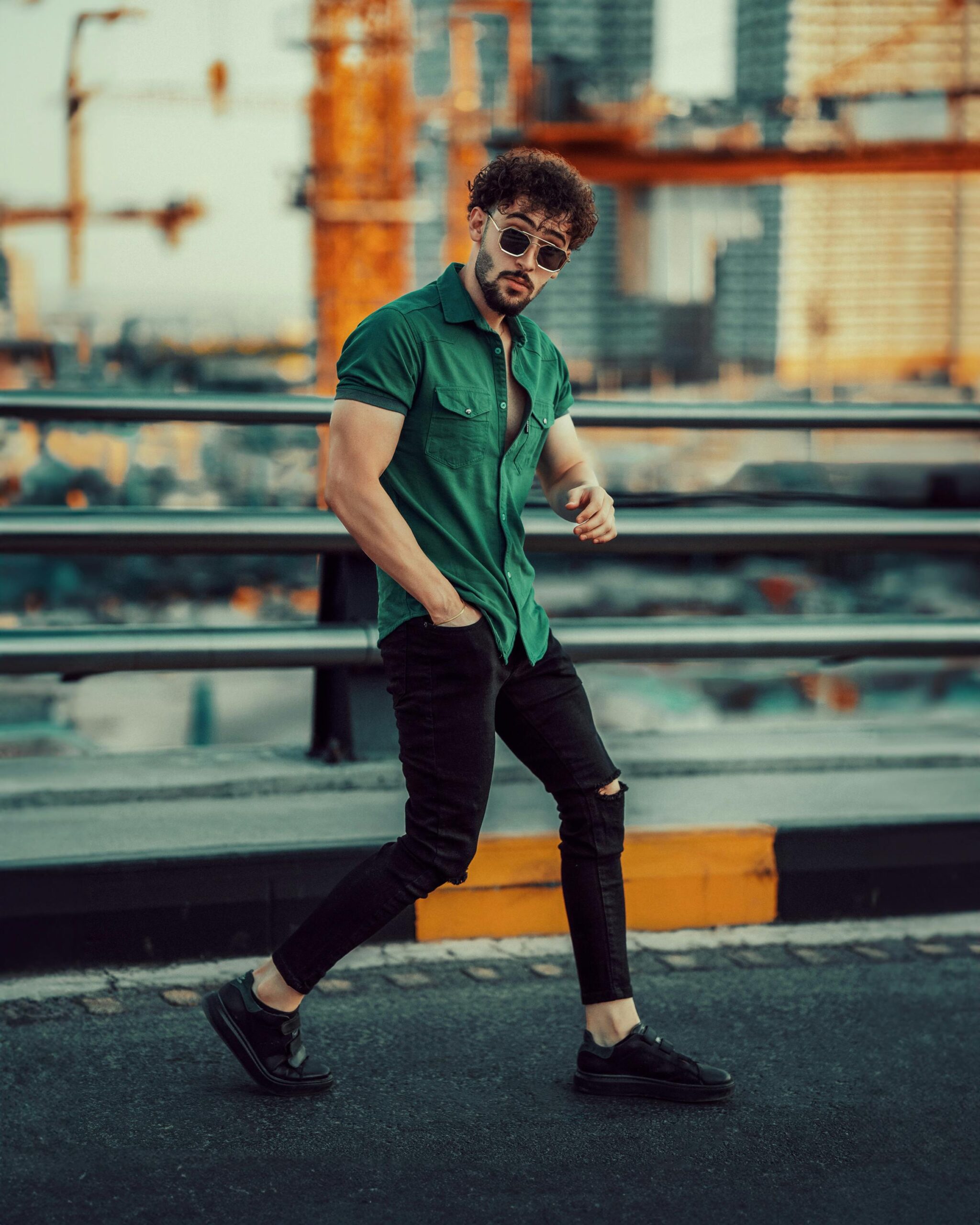 Stylish man with sunglasses in green shirt and ripped jeans posing on bridge against urban backdrop.