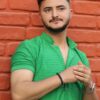 A stylish man in a green shirt poses confidently against a red brick wall.