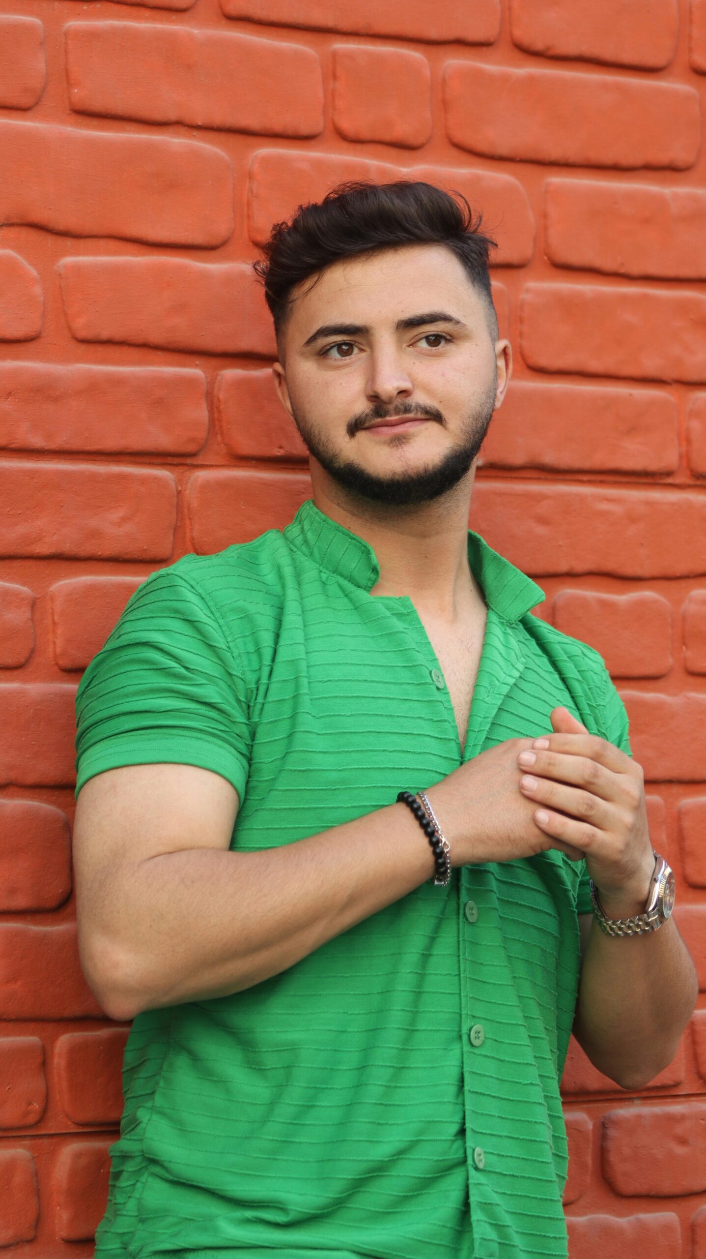 A stylish man in a green shirt poses confidently against a red brick wall.